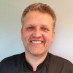 En mann med kort, lysebrunt hår og skjegg smiler til kameraet, iført svart skjorte og stående mot en ensfarget, lys bakgrunn.