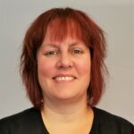 En person med kort, rett, rødbrunt hår og svart skjorte smiler til kameraet mot en vanlig, lys bakgrunn.