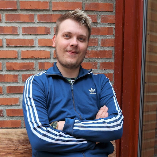 En person med kort, blondt hår og lyst ansiktshår sitter på en trebenk foran en murvegg, iført blå Adidas-jakke og armene i kors.