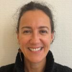 En kvinne med mørkt krøllete hår, iført svart genser, smiler til kameraet foran en lys strukturert vegg.