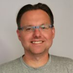 En smilende person med kort, brunt hår, briller og grå t-skjorte står foran en vanlig, lys bakgrunn.