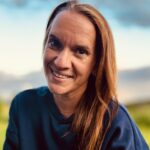 En kvinne med langt brunt hår og mørkeblå topp smiler utendørs, med et uskarpt grønt landskap og skyet himmel i bakgrunnen.
