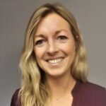 En kvinne med langt, blondt hår smiler mot kameraet. Hun har på seg en mørk topp og står foran en ensfarget, lys bakgrunn.