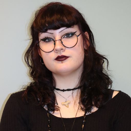 Person med mørkt bølget hår, runde briller, eyeliner, svart leppestift og piercinger i ansiktet, iført svart topp og halskjeder i flere lag, sitter mot en lys bakgrunn.