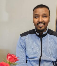 En mann i lyseblå jakke sitter ved et bord med en rød rose i forgrunnen og kjøkkenredskaper på hyller i bakgrunnen.