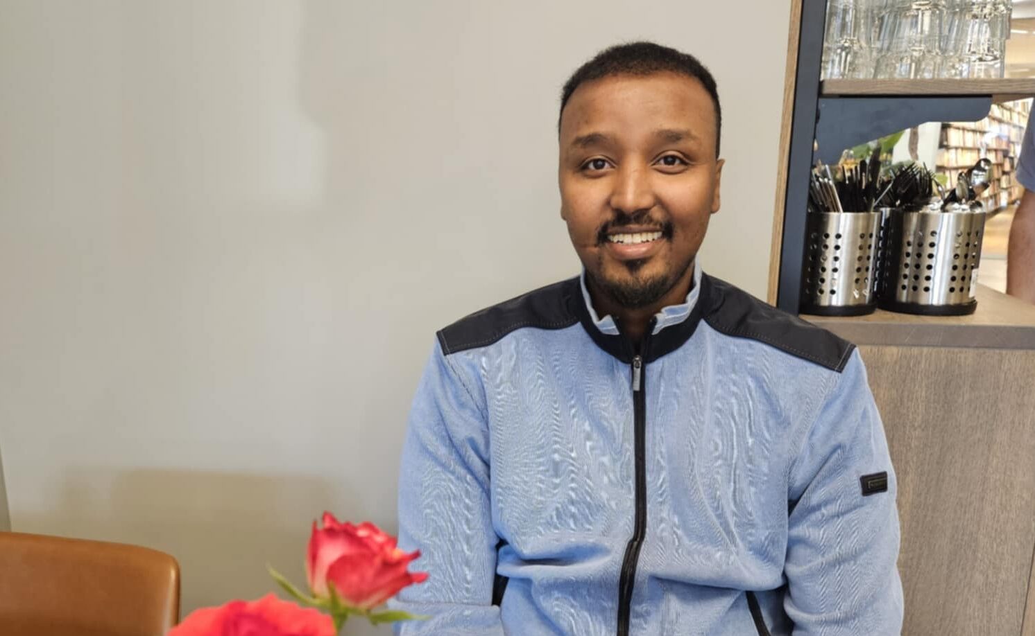 En mann i lyseblå jakke sitter ved et bord med en rød rose i forgrunnen og kjøkkenredskaper på hyller i bakgrunnen.