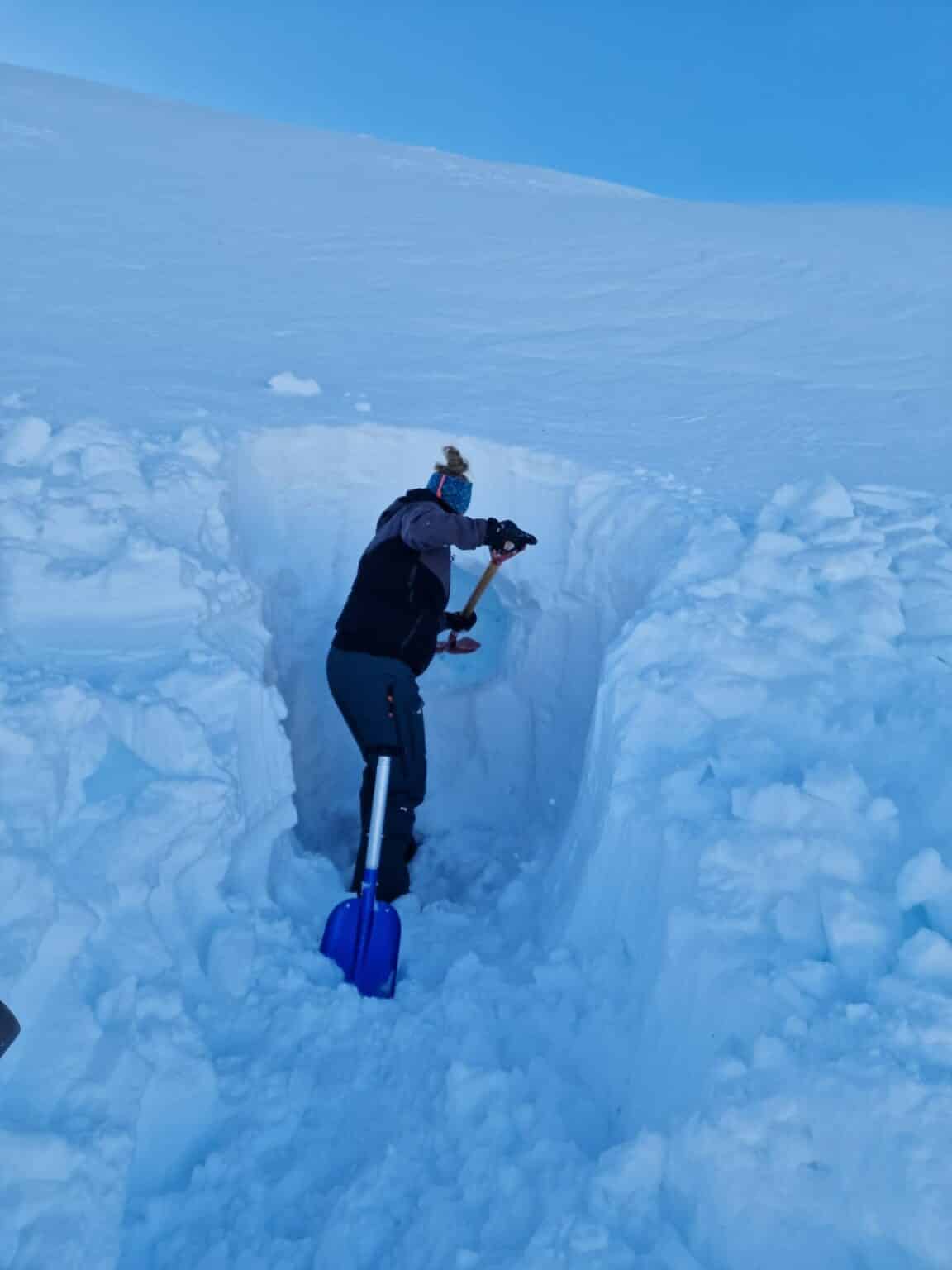 En person står i en dyp grøft gravd ned i snøen og bruker et verktøy til å undersøke eller skjære i snøveggen, mens en blå spade står lent ved siden av.