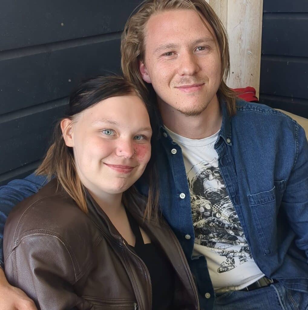En kvinne og en mann sitter tett sammen, begge smiler mot kameraet. Kvinnen har på seg en brun jakke, og mannen har på seg en blå skjorte over en t-skjorte med trykk.