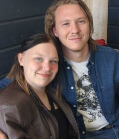 En kvinne og en mann sitter tett sammen, begge smiler mot kameraet. Kvinnen har på seg en brun jakke, og mannen har på seg en blå skjorte over en t-skjorte med trykk.