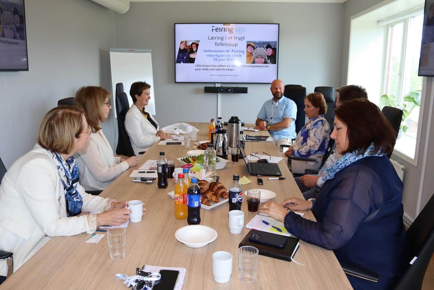 Åtte personer sitter rundt et konferansebord med drikke, kaker og notatbøker og har et møte. En skjerm i bakgrunnen viser en velkomstmelding på norsk.