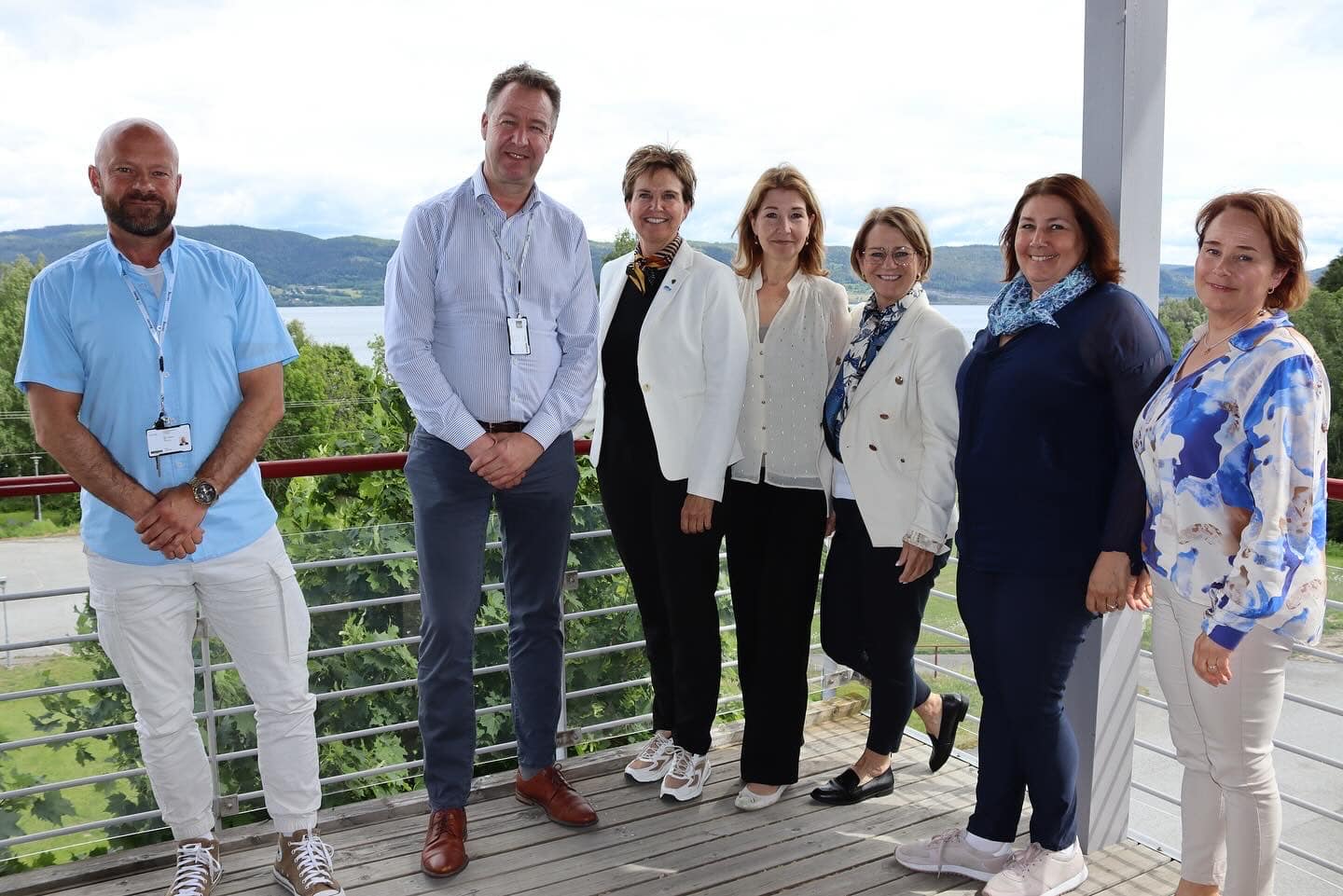 Sju voksne står side om side på en balkong med en naturskjønn innsjø og fjell i bakgrunnen, kledd i uformelle forretningsklær.