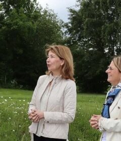 Fire voksne står og snakker utendørs på en gresslette med trær i bakgrunnen; tre kvinner er iført jakker, og en mann har på seg en lyseblå skjorte med nøkkelbånd.