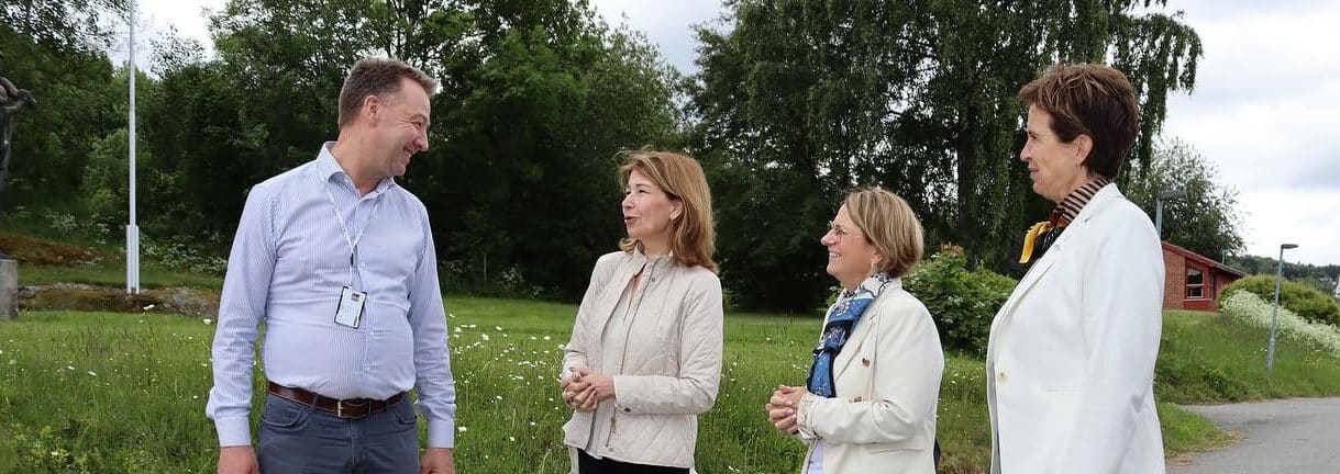 Fire voksne står og snakker utendørs på en gresslette med trær i bakgrunnen; tre kvinner er iført jakker, og en mann har på seg en lyseblå skjorte med nøkkelbånd.