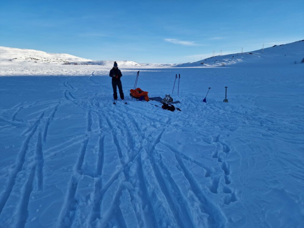 En person står i et snødekt landskap i nærheten av spredt utstyr og skistaver under en klarblå himmel, med snødekte åser i bakgrunnen.