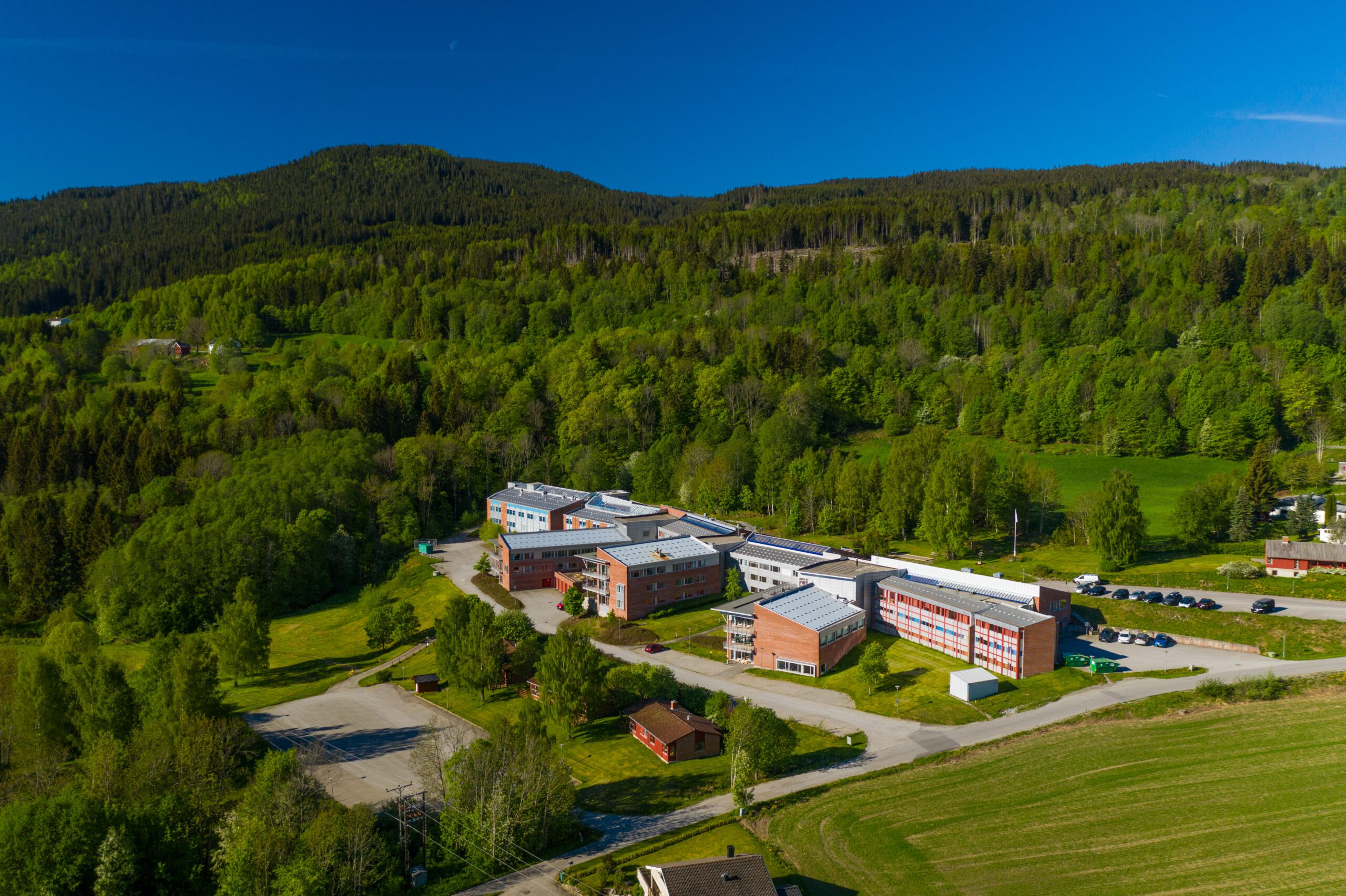 Flyfoto av en moderne skolecampus med flere mursteinsbygninger omgitt av grønne trær og åkrer, mot en skogkledd åsside under en klar blå himmel.