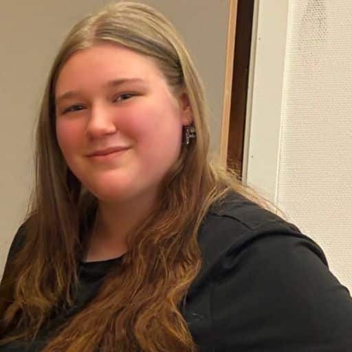 En person med langt, lysebrunt hår og svart topp står innendørs, vendt mot kameraet og smiler lett.