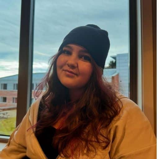 En person med langt, brunt hår, svart lue og lys jakke sitter innendørs foran et vindu med bygninger og en overskyet himmel utenfor.