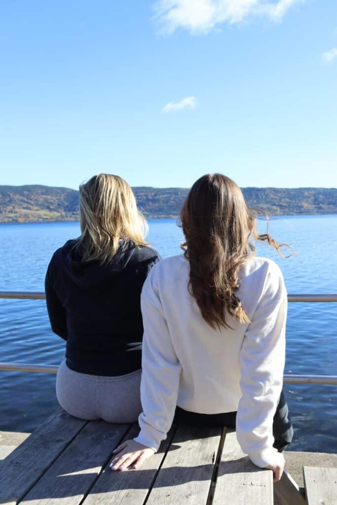 To personer sitter på en trebrygge med utsikt over en rolig innsjø med åser i bakgrunnen under en klarblå himmel.
