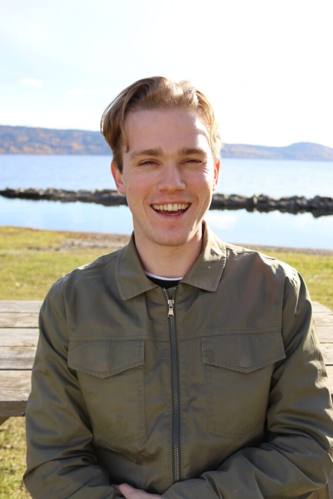 En ung mann i grønn jakke smiler og sitter ved et trebord utendørs, med en innsjø og fjerne åser i bakgrunnen.