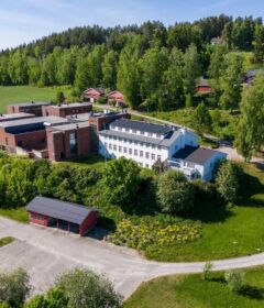 Flyfoto av et stort bygningskompleks med røde og hvite strukturer, omgitt av trær, gress og noen små uthus i et landlig område.