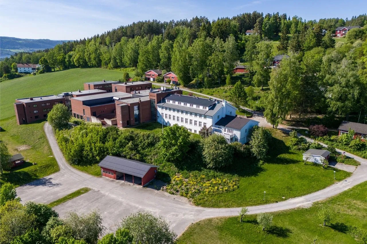 Flyfoto av et stort bygningskompleks med røde og hvite strukturer, omgitt av trær, gress og noen små uthus i et landlig område.