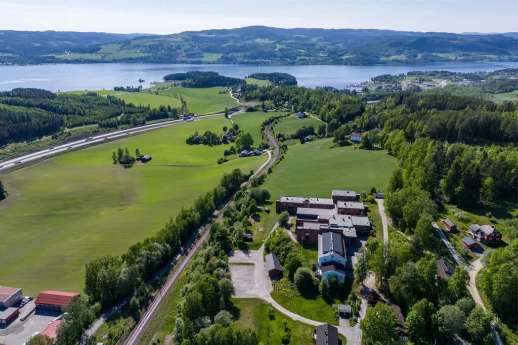 Flyfoto av et ruralt landskap med tett bebyggelse, jernbane, grønne åkre, skogsområder og en innsjø i bakgrunnen under en klar himmel.