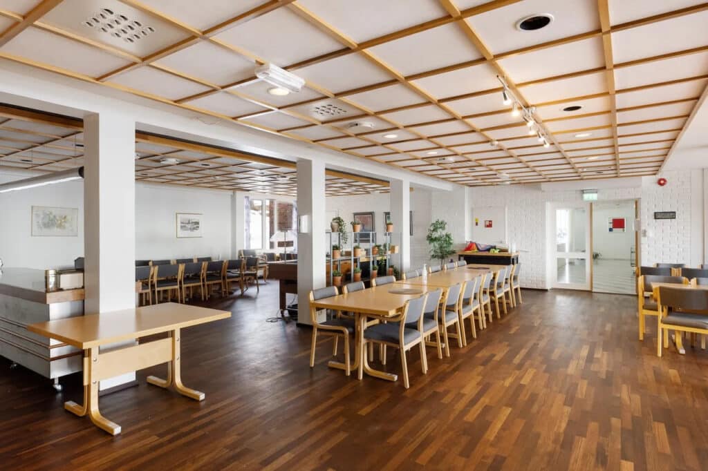 En romslig spiseplass med tregulv, bord og stoler, et tak med rutenettmønster og store vinduer som slipper inn naturlig lys.