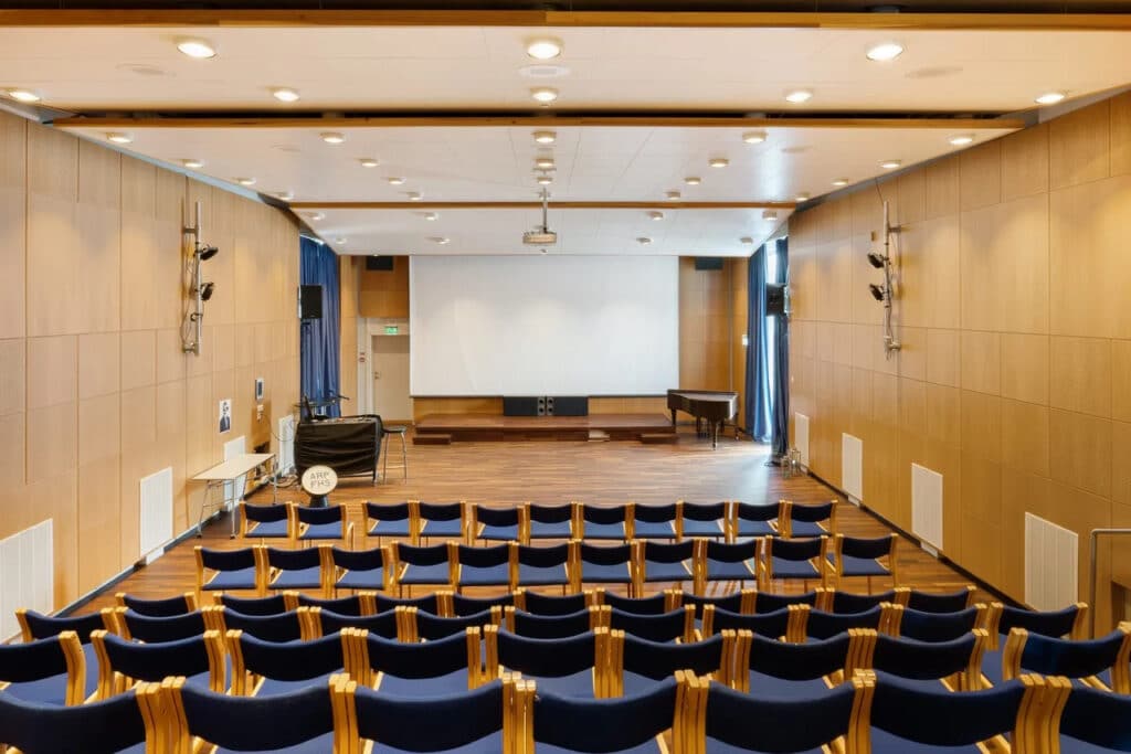 Rader av tomme, blå stoler står foran en scene med et stort, hvitt lerret, et piano og trepanel på veggene i et godt opplyst auditorium.