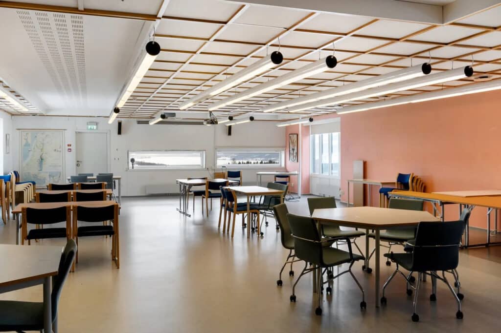 Et romslig klasserom med runde og rektangulære bord, diverse stoler, store vinduer og rosa vegger, med kart og kunstverk utstilt.