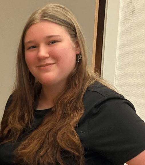 Skjermbilde 2026-03-03 121534 En ung kvinne med langt brunt hår og svart skjorte smiler mens hun står innendørs ved en vegg.