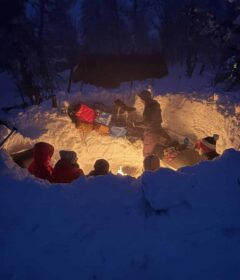 En gruppe mennesker sitter rundt et bål i en snødekt grop om natten, omgitt av snøbanker og vinterutstyr, med trær og et gapahuk i bakgrunnen.
