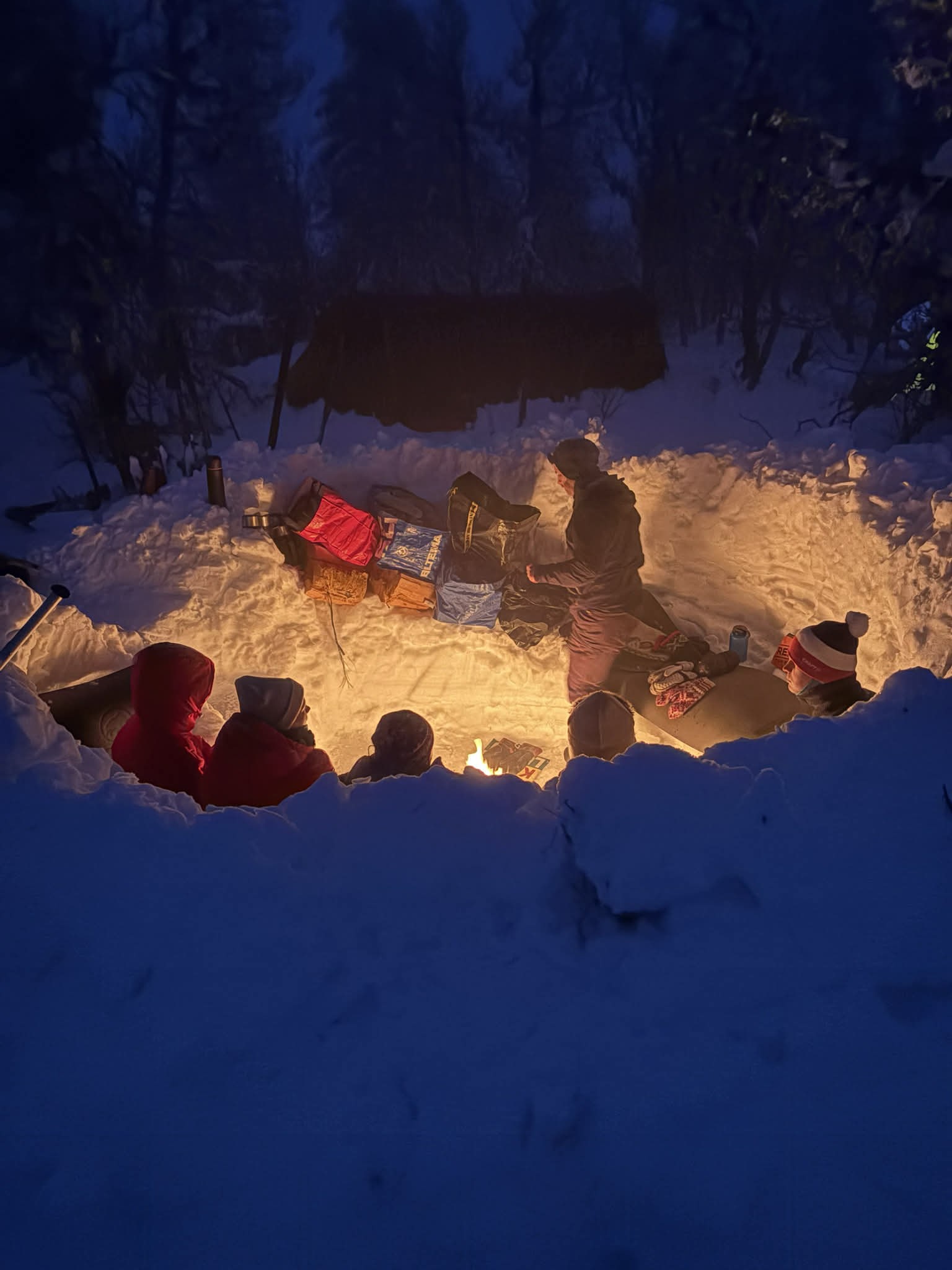 En gruppe mennesker sitter rundt et bål i en snødekt grop om natten, omgitt av snøbanker og vinterutstyr, med trær og et gapahuk i bakgrunnen.