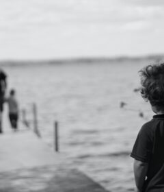 Et barn står ved et tre og ser på to personer som går på en brygge som strekker seg ut i en innsjø; bildet er i svart-hvitt.